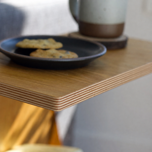 A close-up of an Alden table in oak with layered plywood edges, holding a black plate with cookies and a ceramic mug on a coaster in a softly lit indoor setting.