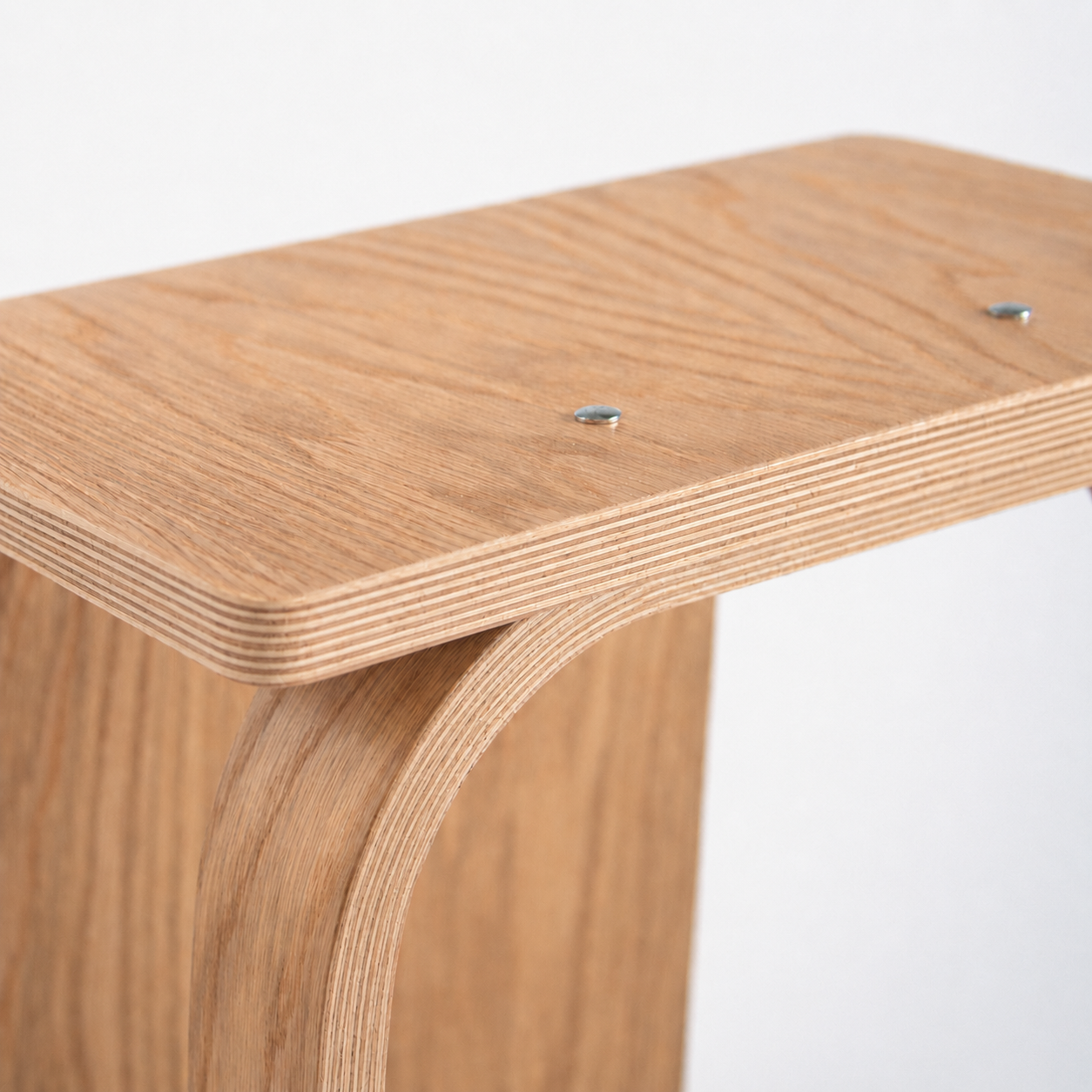 Close-up of a light natural wood stool showing the layered plywood construction, a curved front support, and two visible metal screws on the seat against a plain white background.
