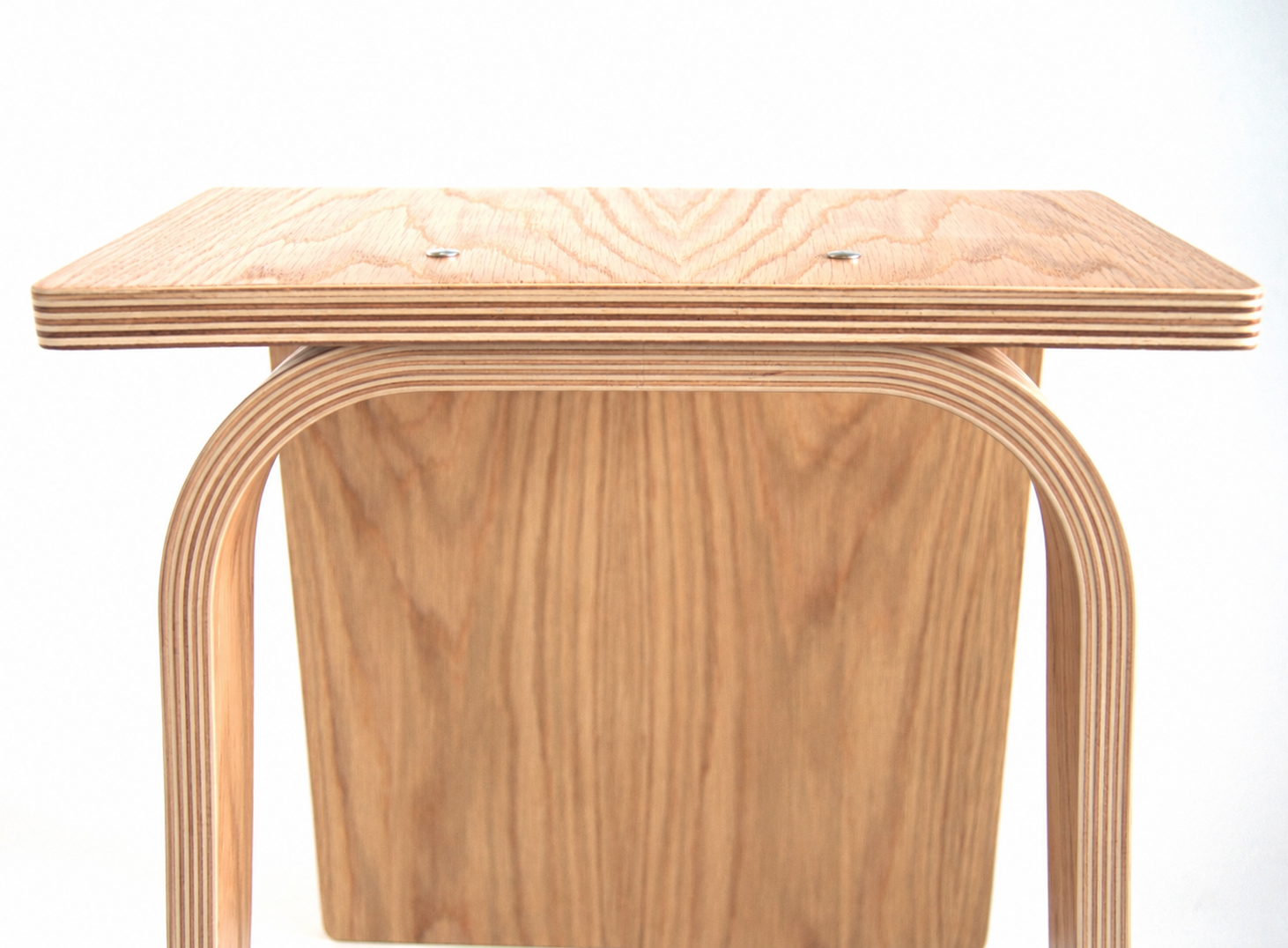 A close-up of a light natural oak wood stool with a square seat and curved legs, showing the layered plywood construction against a white background.