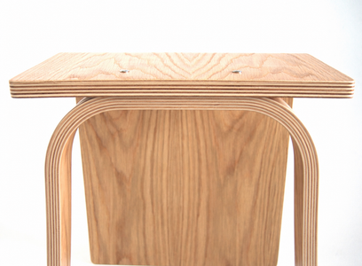 A close-up of a light natural oak wood stool with a square seat and curved legs, showing the layered plywood construction against a white background.