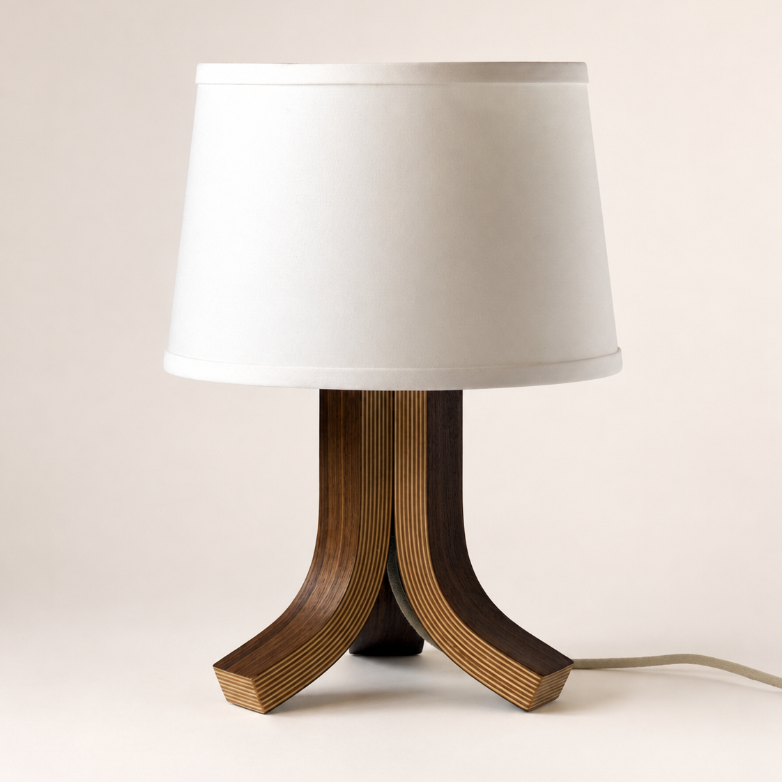 A modern table lamp with a white fabric drum shade and a sculptural three-legged base made of layered wood veneer in dark and medium brown tones, shown against a plain light beige background.