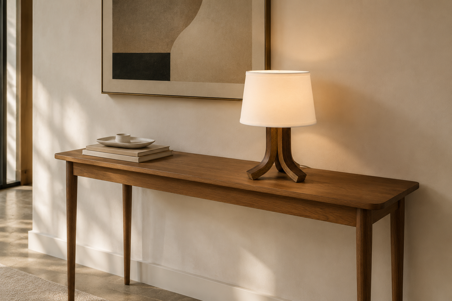 A tripod-base table lamp with a white shade lighting a warm-toned wooden console table in a styled interior setting.