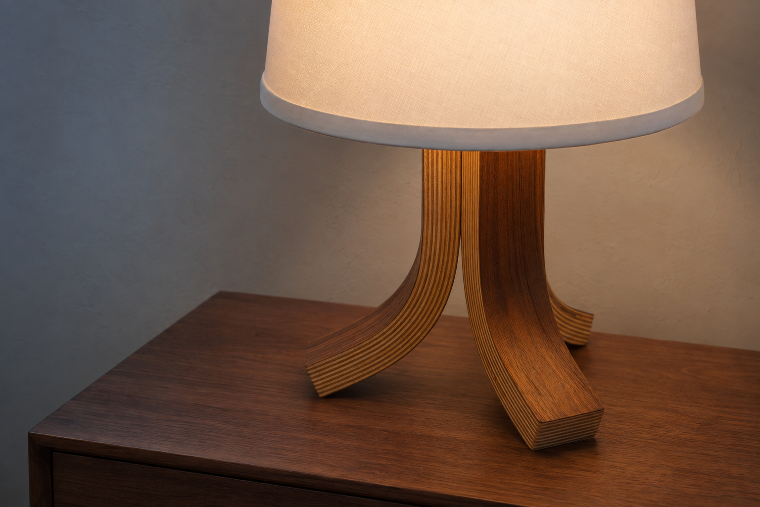 A table lamp with a warm white fabric drum shade and a sculptural layered wood base, shown lit on a wooden side table against a neutral gray wall.