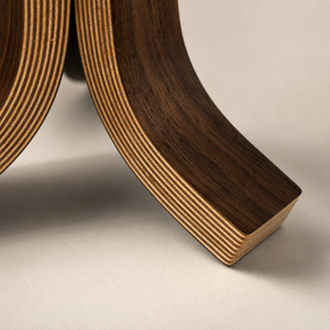 Close-up of a curved wooden lamp base showing layered plywood construction in dark brown and light tan tones, set against a neutral light gray background.