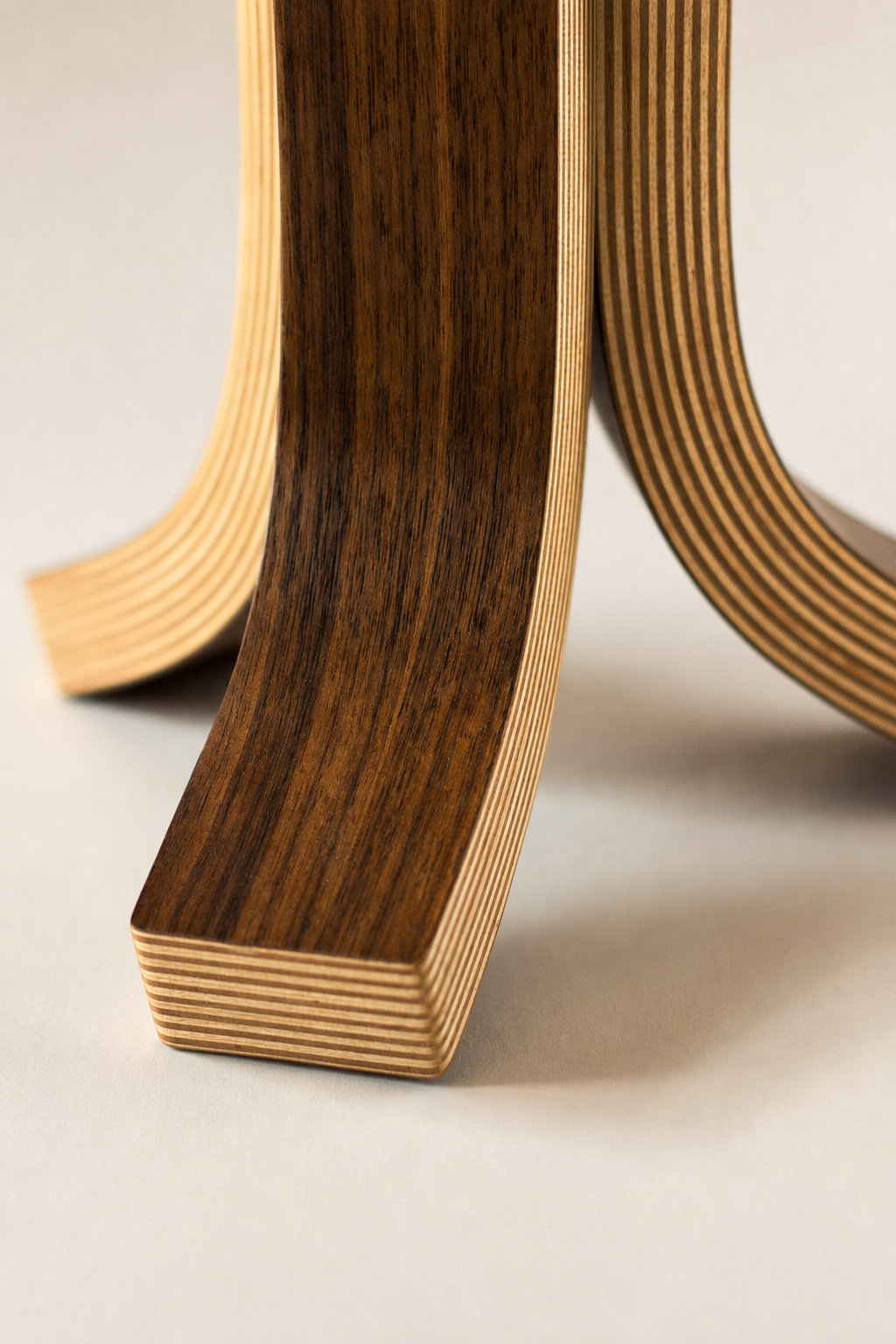 Close-up of a curved wooden lamp base with layered plywood construction, showing a walnut wood veneer and light natural edges, on a neutral light beige background.