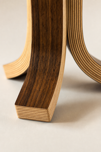 Close-up of a curved wooden lamp base with layered plywood construction, showing a walnut wood veneer and light natural edges, on a neutral light beige background.